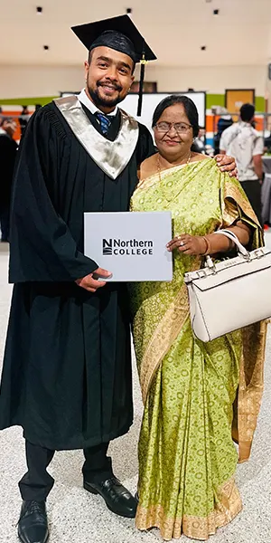A graduate in cap and gown holding a college folder, standing with a family member at their convocation