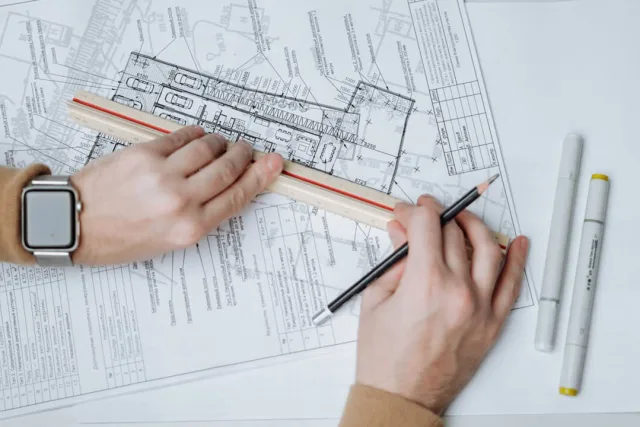 Two hands drawing on a floor plan with a scale rule and a pencil
