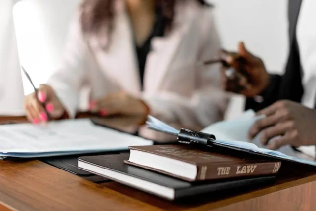 A bound volume lettered “The Law” on a desk, with two people signing papers behind it