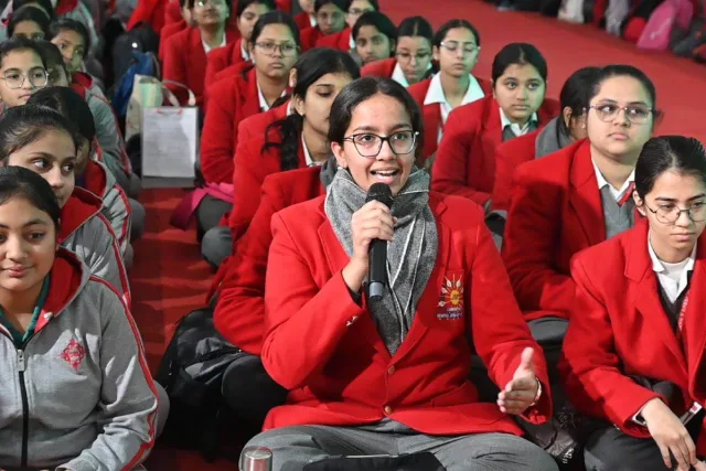 A student asking a question into a microphone from the floor of the hall, with classmates seated around her.